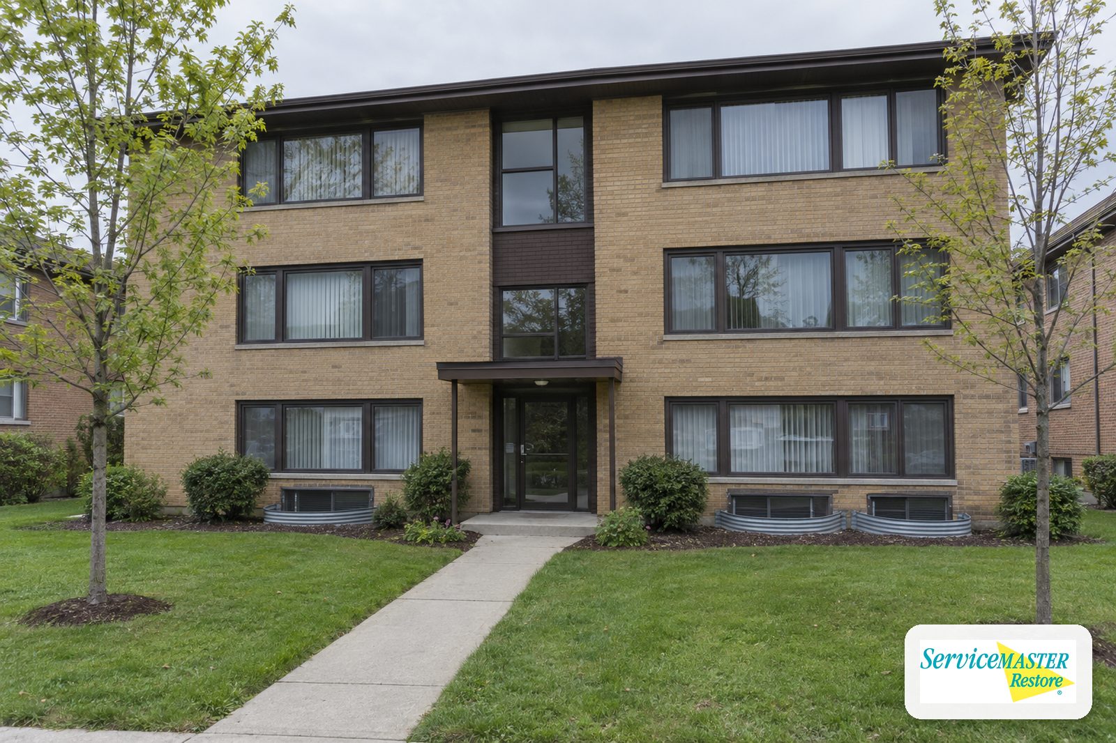 Three-story 1970s tan-brick garden apartment building with window wells along the garden level
